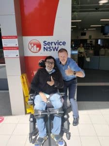 Person sitying in power wheelchair in front of Service NSW with driving instructor next to him, both doing thumbs up after passing driving test.