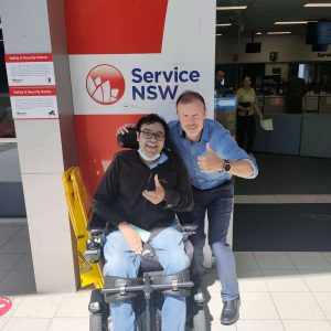 Person sitying in power wheelchair in front of Service NSW with driving instructor next to him, both doing thumbs up after passing driving test.