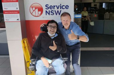 Person sitying in power wheelchair in front of Service NSW with driving instructor next to him, both doing thumbs up after passing driving test.